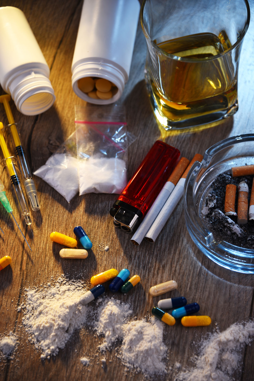 Verschiedene Drogenutensilien auf einem Tisch: Tabletten, Spritzen, Drogenpulver, Alkohol und Zigaretten, symbolisch für Alkohol- und Drogenvergehen bei der MPU.